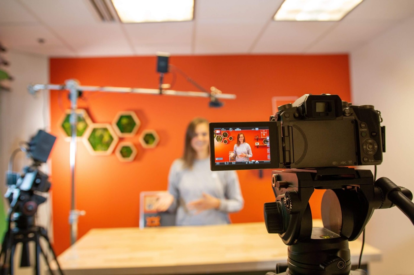 A video camera in the foreground is capturing an image of a woman standing in front of an orange studio wall